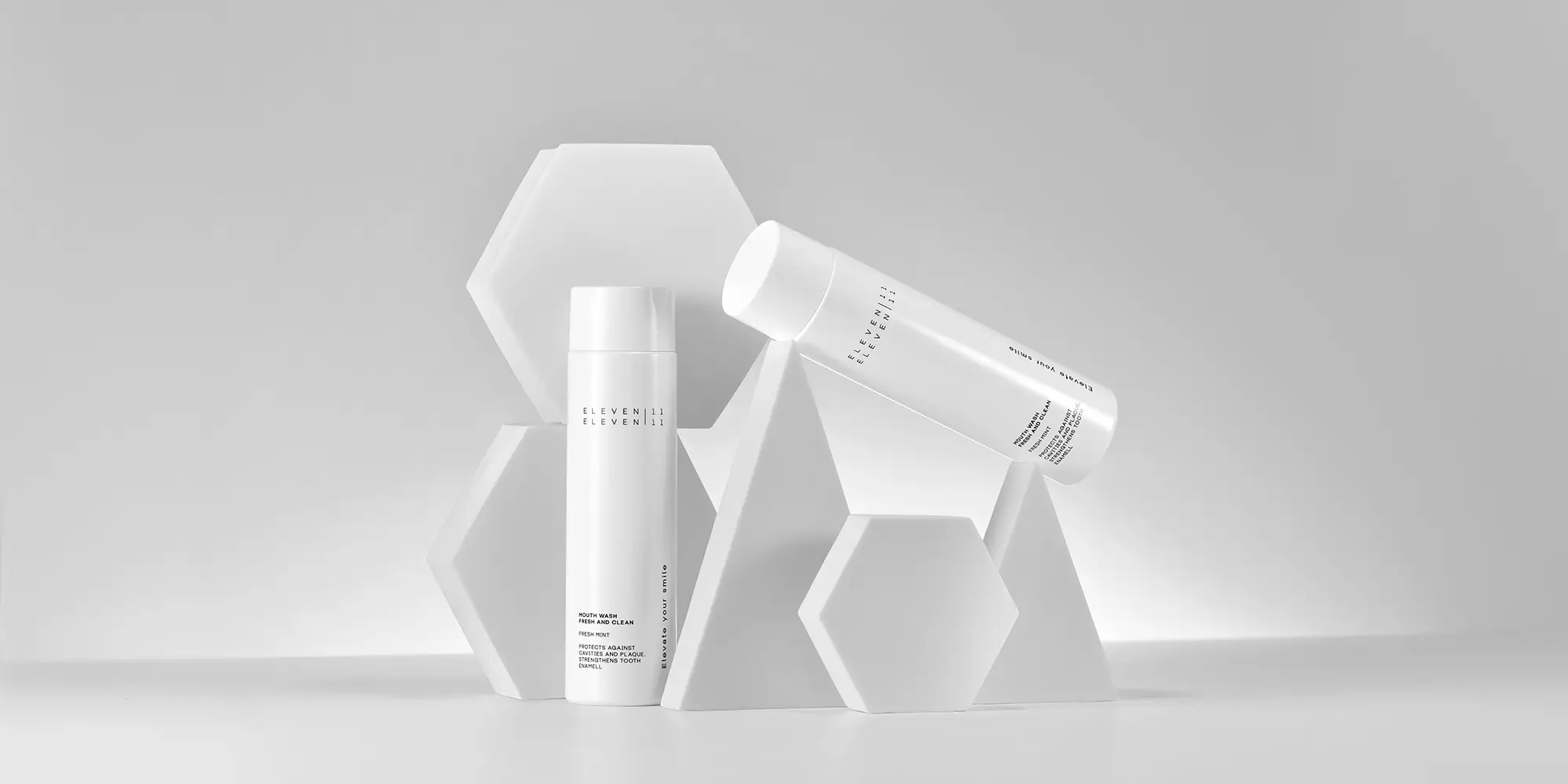 Zwei weiße Mundspülungsflaschen der Marke ELEVEN ELEVEN stehen und liegen auf weißen geometrischen Formen wie Pyramiden und Sechsecken. Die Komposition wirkt architektonisch und modern.