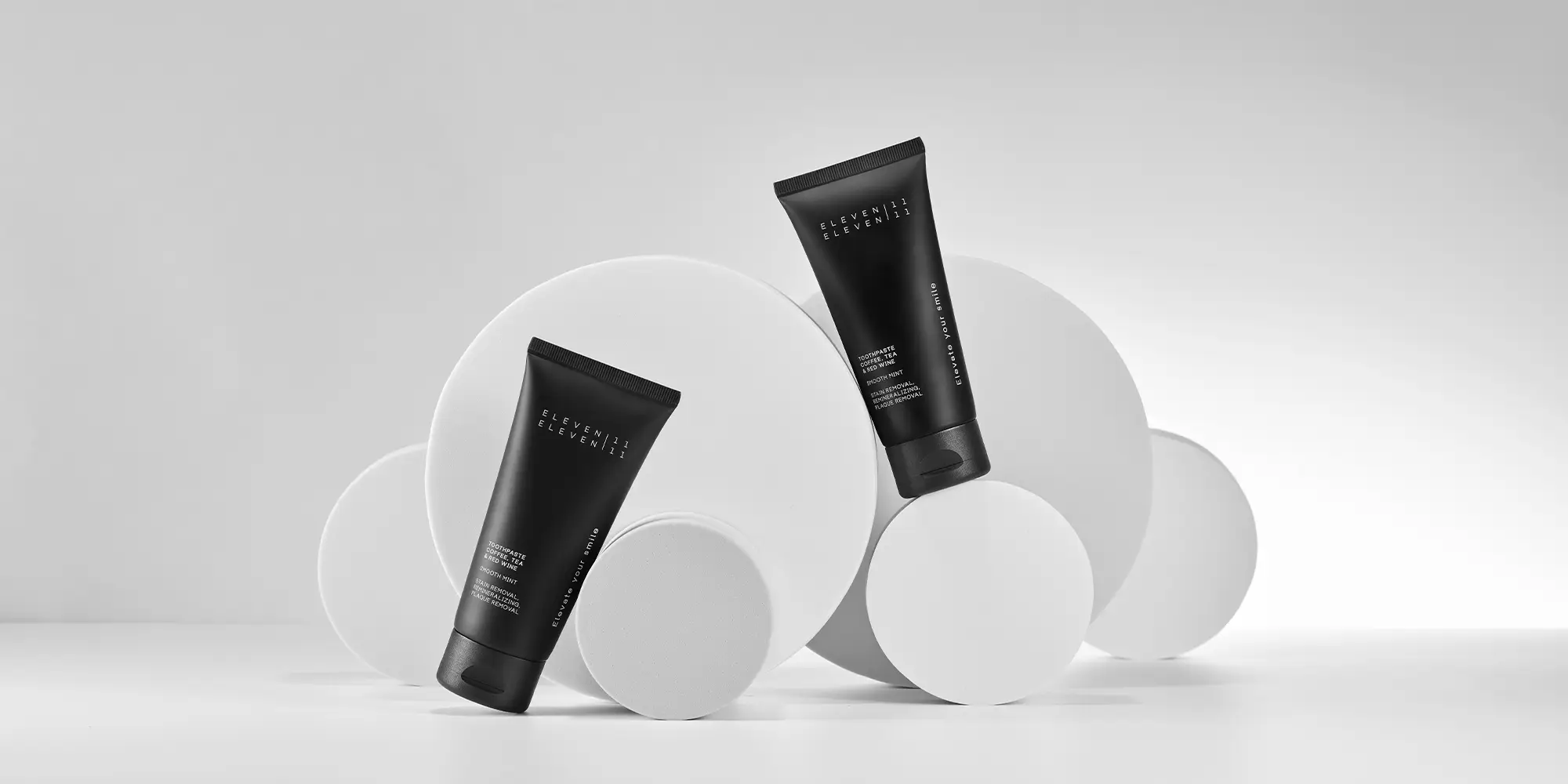 Zwei schwarze Zahnpasta-Tuben der Marke ELEVEN ELEVEN lehnen an runden, weißen Formen. Die Anordnung wirkt kunstvoll und designorientiert, mit dem Fokus auf die stilvolle Produktverpackung.