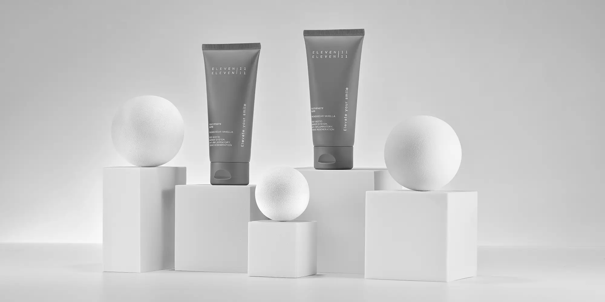 Zwei graue Zahnpasta-Tuben der Marke ELEVEN ELEVEN stehen auf unterschiedlich hohen, weißen Sockeln. Runde, weiße Kugeln ergänzen die stilvolle Präsentation.