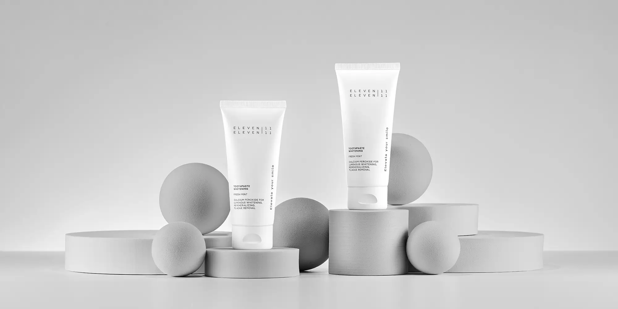 Zwei weiße Zahnpasta-Tuben der Whitening-Serie stehen auf unterschiedlich hohen, zylindrischen Podesten. Runde Kugeln umgeben die Produkte und schaffen ein harmonisches, ästhetisches Gesamtbild.