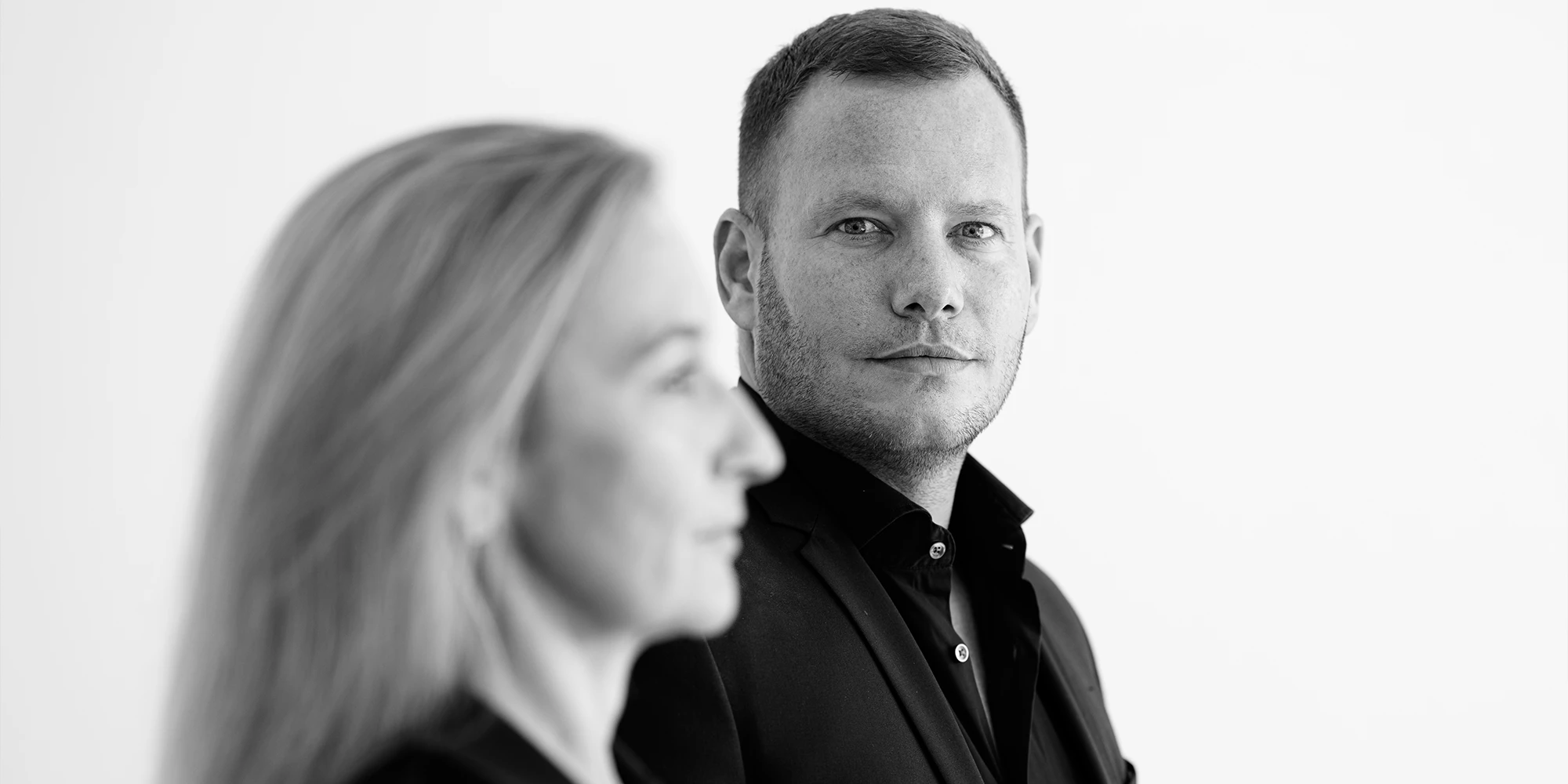 Das Bild zeigt eine Person in Nahaufnahme, in Schwarz-Weiß. Der Fokus liegt auf einem Mann mit kurzem Haar und leichtem Bart, der direkt in die Kamera blickt. Er trägt ein dunkles Hemd und möglicherweise ein Jackett. Der Hintergrund ist hell und schlicht gehalten. Der Gesichtsausdruck wirkt ruhig, professionell und selbstbewusst. Es handelt sich um dr. med. dent. (D.M.D.) Oliver Spitz. Im Vordergrund erkennt man etwas unscharf die Silhouette einer Frau mit blonden Haaren. Hierbei handelt es sich um Bianca Paulibl, die Business Managerin von ELEVEN ELEVEN.